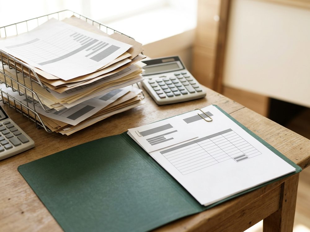 Stacks of mismatched timesheet forms in wire trays beside a calculator, with one finished invoice and its stapled summary sheet in a green folder